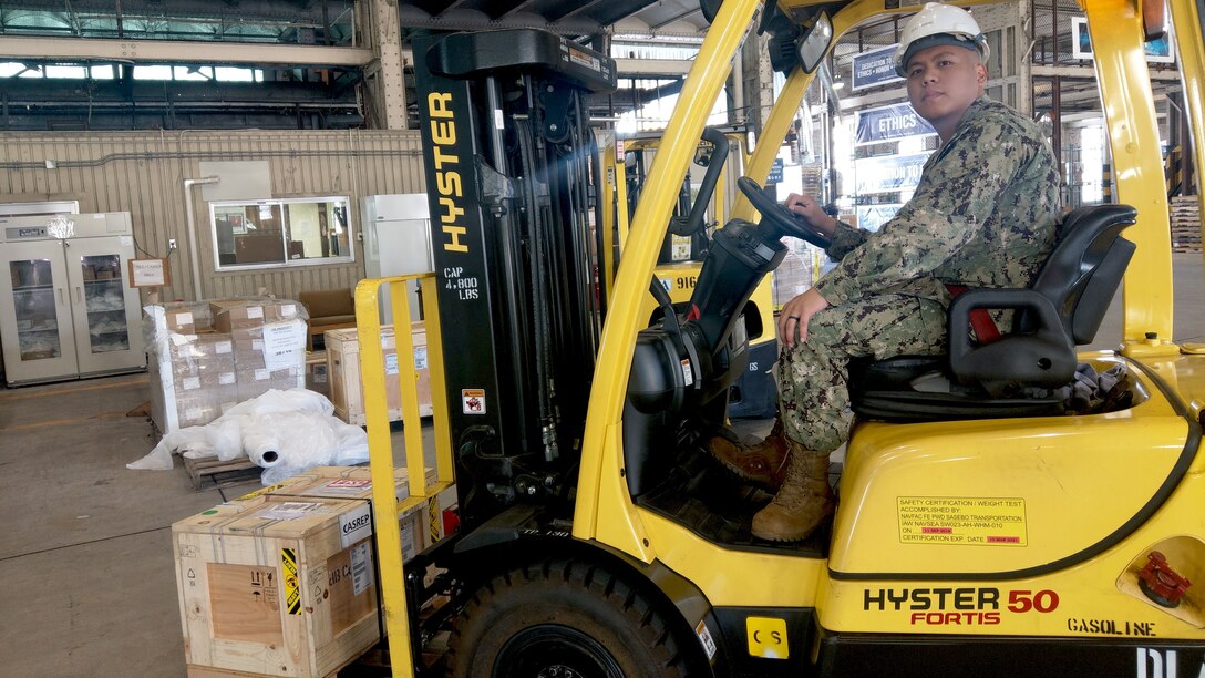 A sailor drives a forklift carrying boxes of supplies in a warehouse