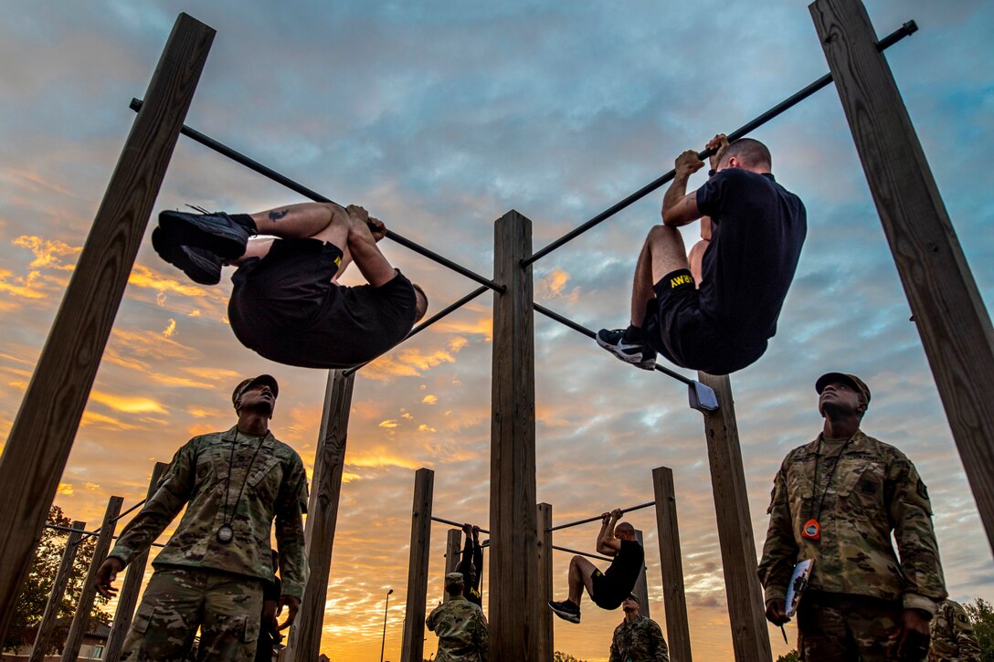 Soldiers hanging from high bars tuck their legs as others observe from the ground.