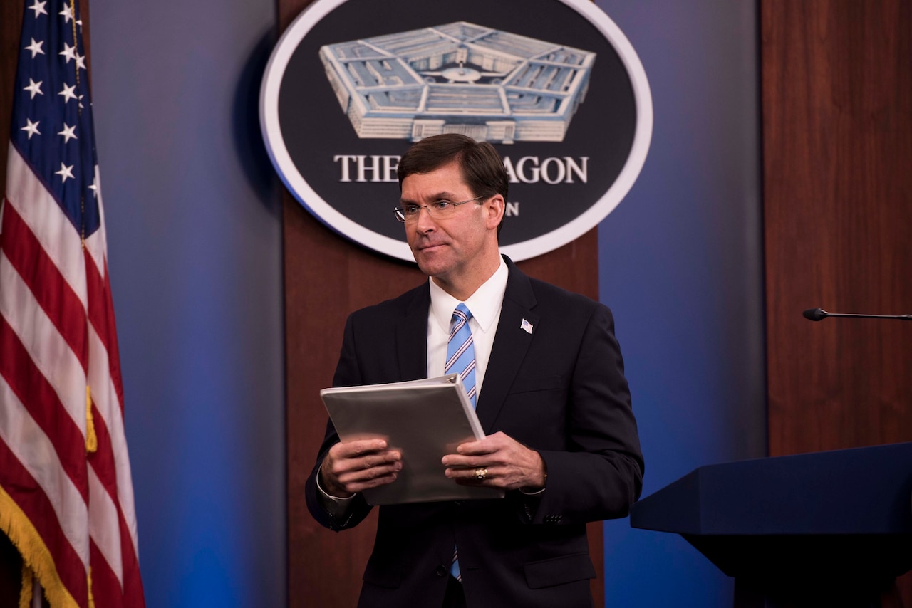 Man in suit holds a white binder as he walks away from a lectern.