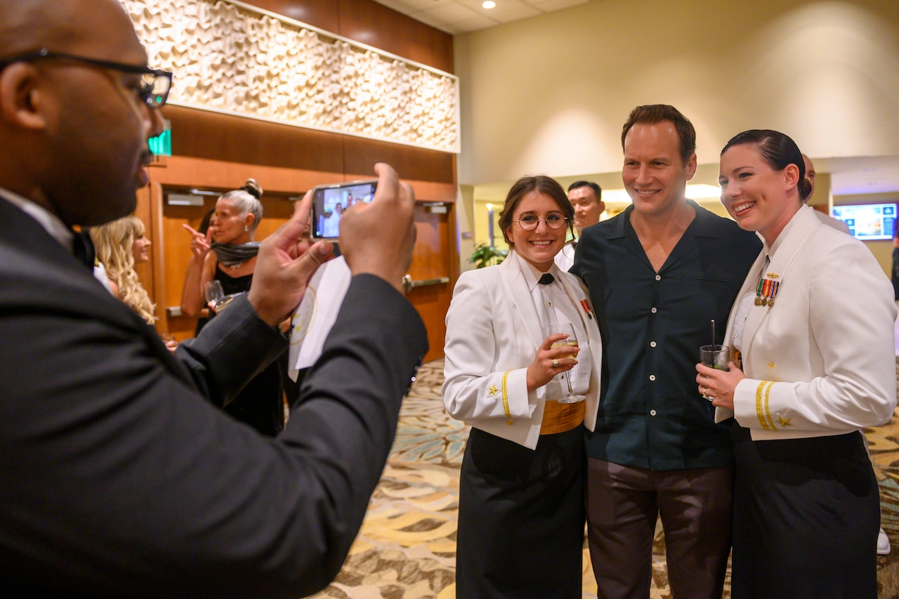 A man takes a cellphone photo of actor Patrick Wilson standing between two female sailors in dress uniform at a gala.