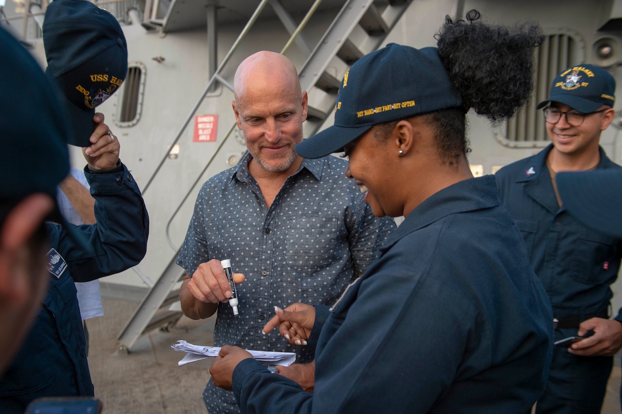 Actor Woody Harrelson signs an autograph for a female sailor as other sailors smile beside her.