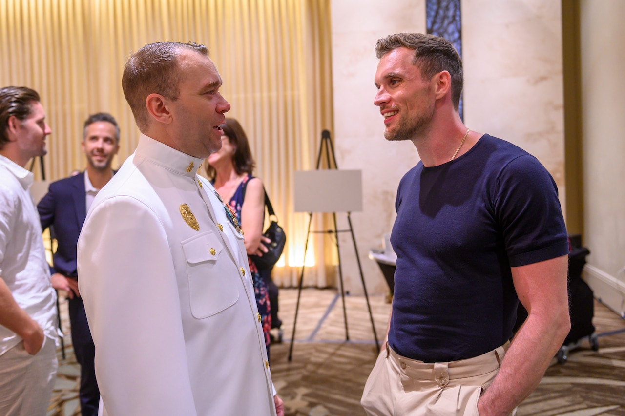 A Navy sailor in dress white uniform talks with actor Ed Skrein. Three others can be seen chatting in the background.
