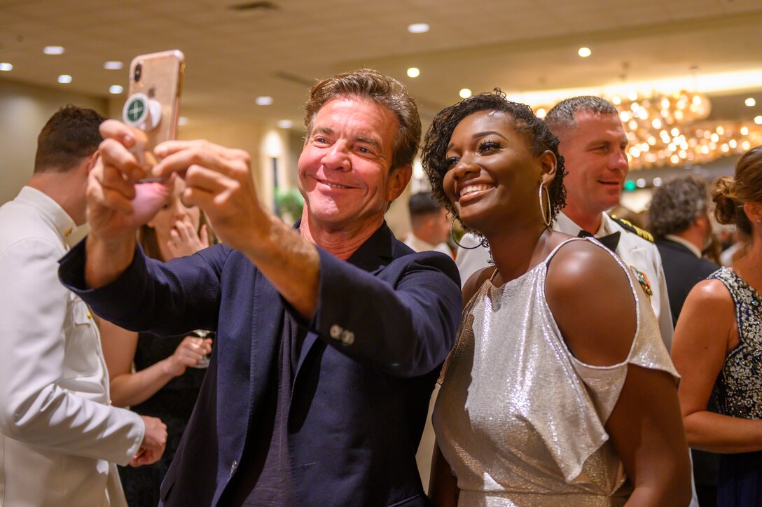 Actor Dennis Quaid holds out a cellphone to take a selfie with a woman wearing a fancy dress. Others mill around behind them.