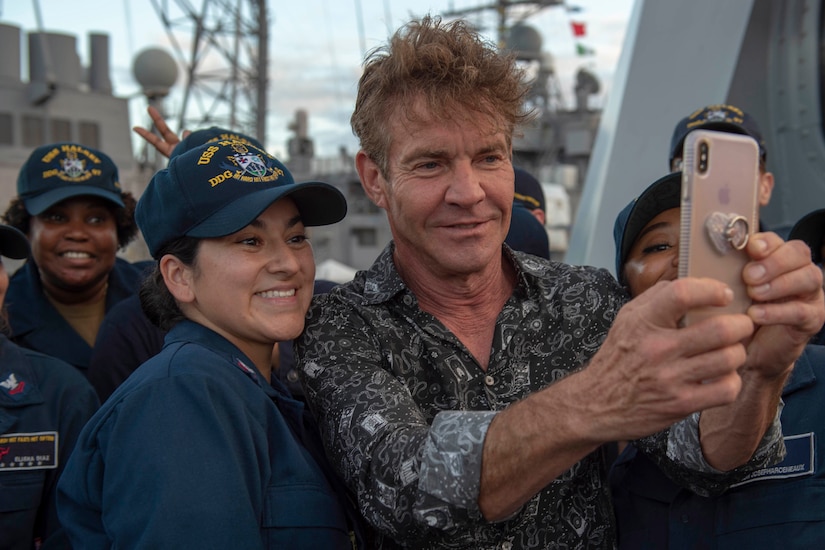 Navy Seaman Aja Bleu Jackson
ALT: Actor Dennis Quaid holds a phone while standing between two female sailors to get a selfie with them on a ship. Others pose in the background.