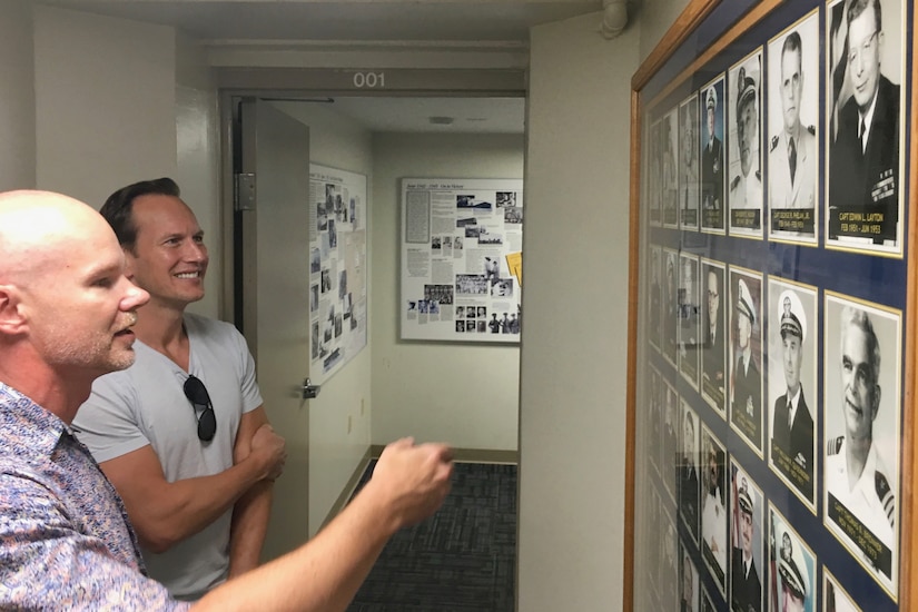 Two men look at framed photographs on a wall.