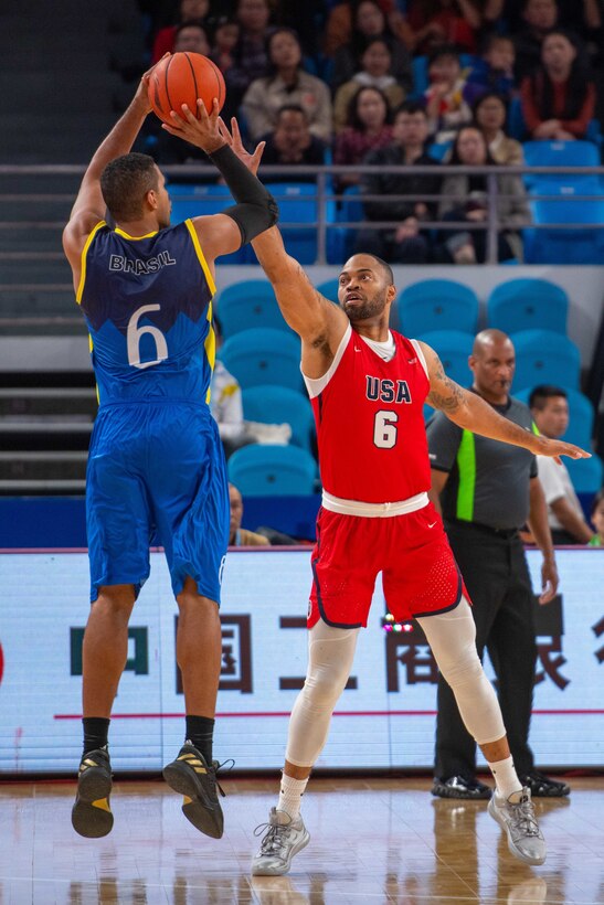 Blue-clad basketball player puts up a jump shot as red-clad opponent tries to block it.