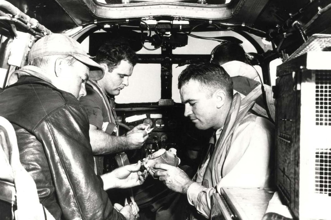 Soldiers sitting in a cockpit wearing World War II-era cold-weather flying clothes hold a pigeon.