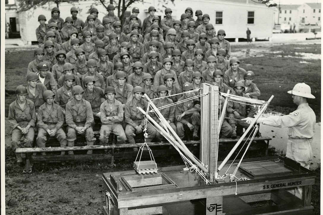 Soldiers sitting in bleachers watch a soldier use a wooden model of a crane to demonstrate how ships are loaded.