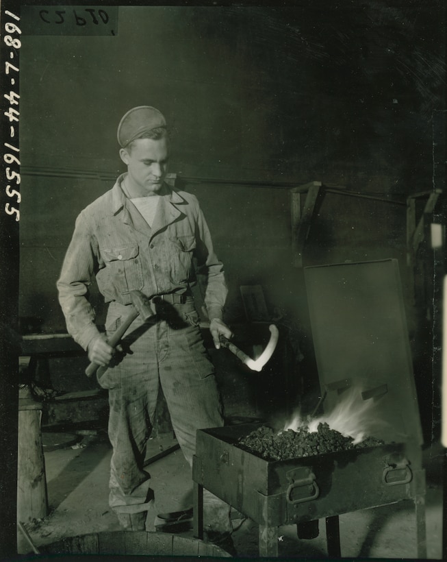 A soldier with a hammer in one hand holds a glowing hot metal bar over coals.