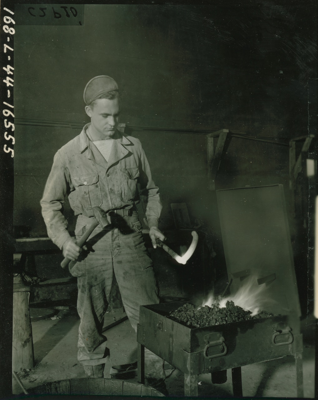 A soldier with a hammer in one hand holds a glowing hot metal bar over coals.