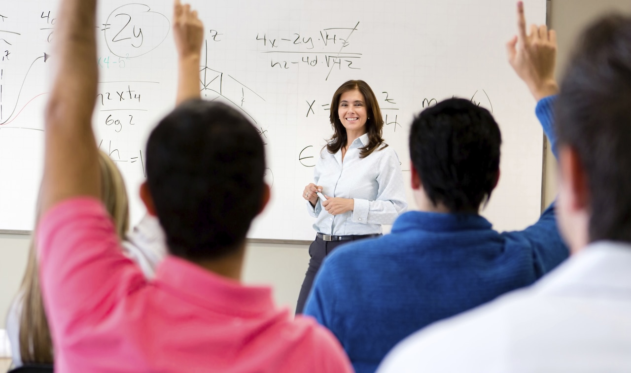 A teacher stands in front of a whiteboard as students raise their hands to answer her question.