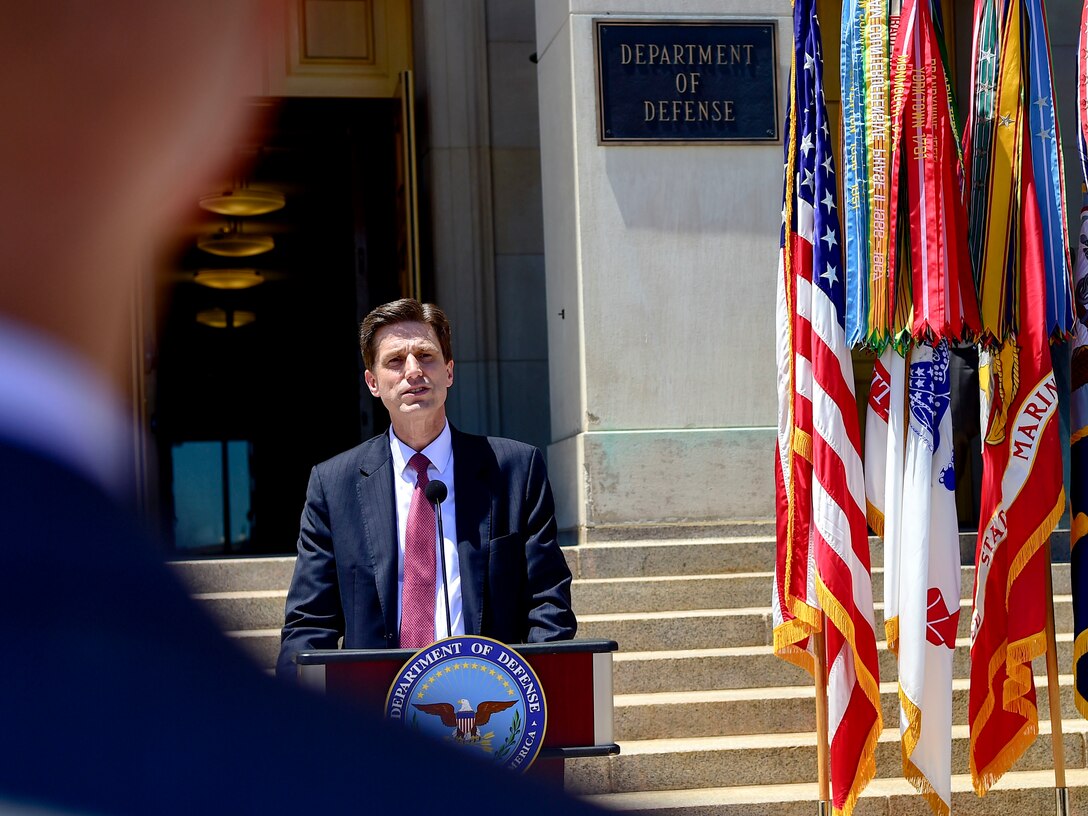 A man speaks into a microphone on a podium that has a Defense Department seal. The U.S. flag is to his right.
