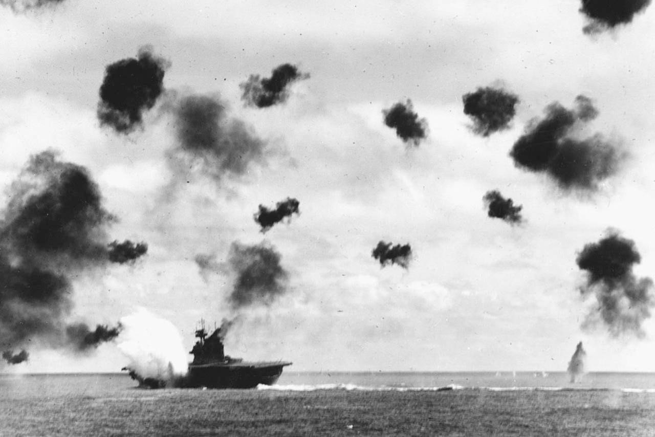 Pufffs of dark smoke dot the sky as bombs fall on a ship.