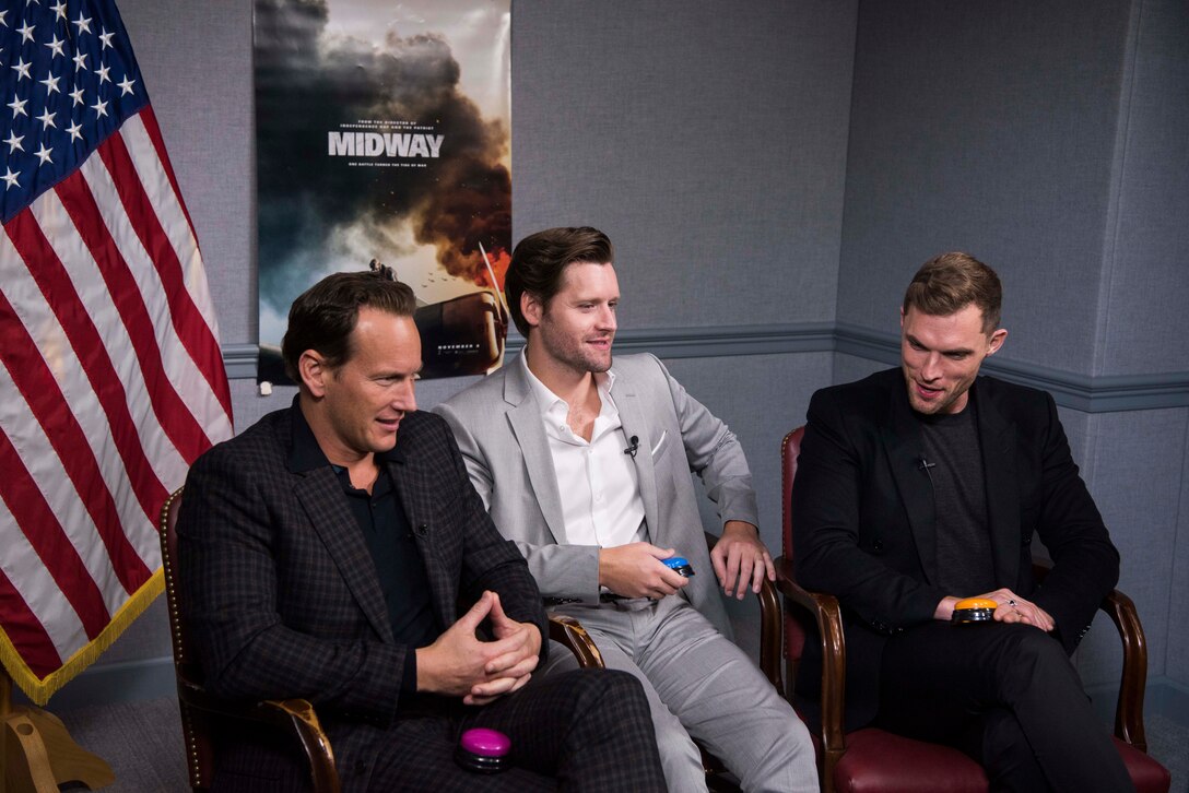 Three men in suits sit in chairs next to one another. In the background is a poster from the movie “Midway.” A U.S. flag is on the left side of the men.
