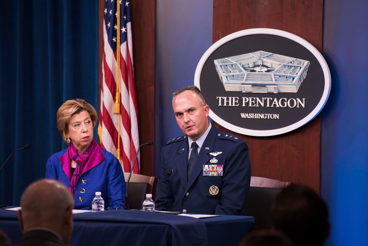 A woman and a man in military uniform sit at a table. Behind them hangs a sign with the words ‘The Pentagon ... Washington.’