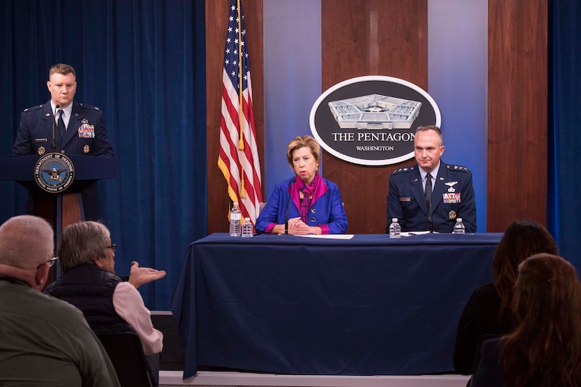A woman and a man in military uniform sit at a table. Behind them hangs a sign with the words ‘The Pentagon ... Washington.’
