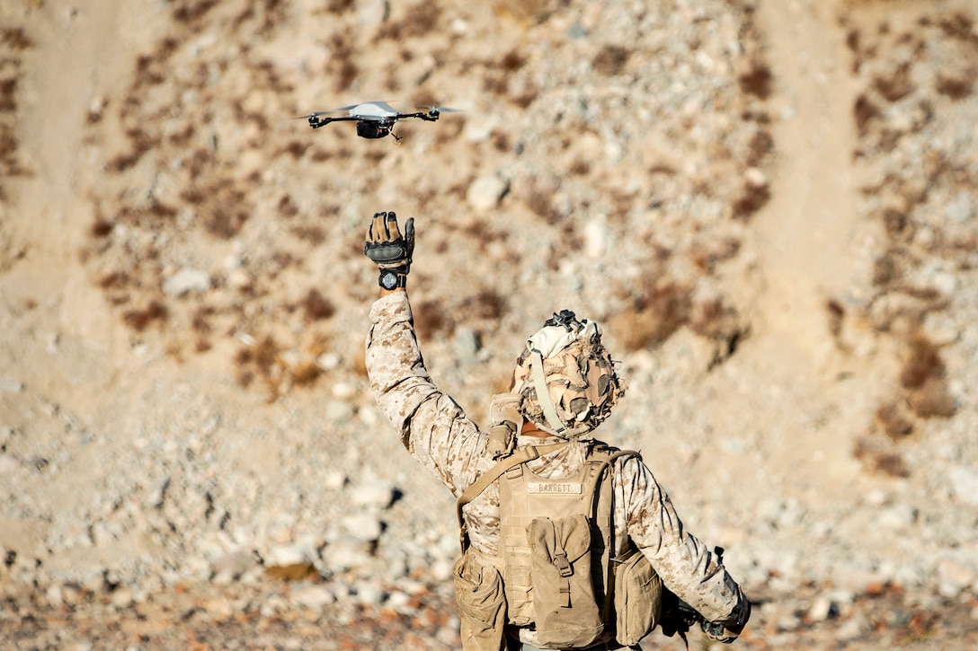 A Marine raises his arm as a drone hovers above it, against a brown desert background.