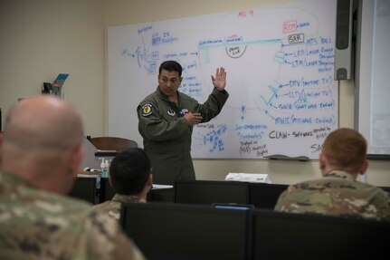 Between the space of three students heads is an instructor standing at the front of a classroom in front of a whiteboard explaining a lesson while utilizing his arms and hands to help emphasize his words.