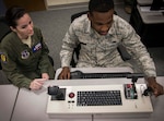 An instructor sits next to a student at a tabletop simulator to help guide him with proper procedures. This simulator is specifically to help train sensor operators. It has a keyboard and three levers.