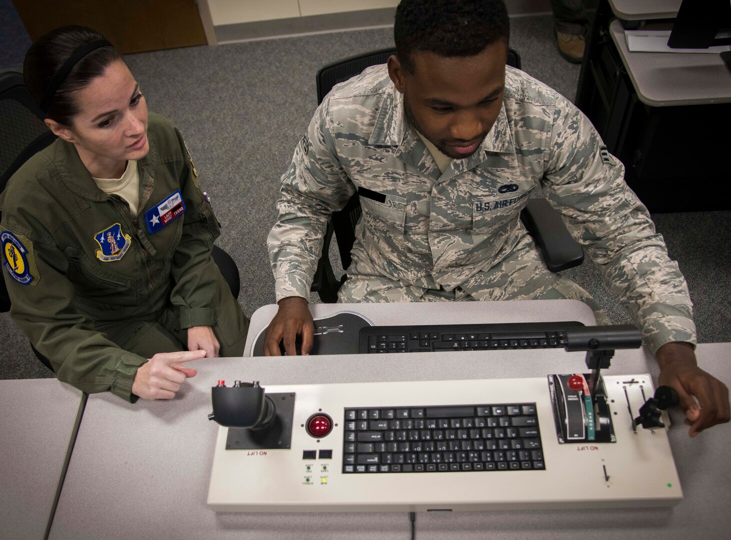 An instructor sits next to a student at a tabletop simulator to help guide him with proper procedures. This simulator is specifically to help train sensor operators. It has a keyboard and three levers.