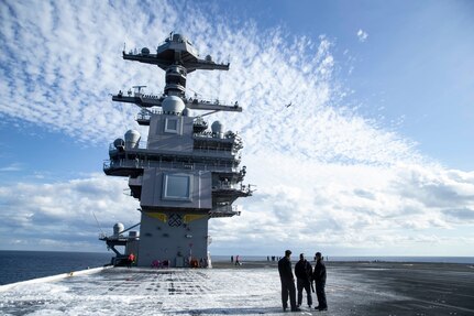 An aircraft flies over USS Gerald R. Ford (CVN 78) during dual-band radar testing while Aqueous Film Forming Foam (AFFF) coats the flight deck following a succussful test of the flight deck AFFF hose reels.