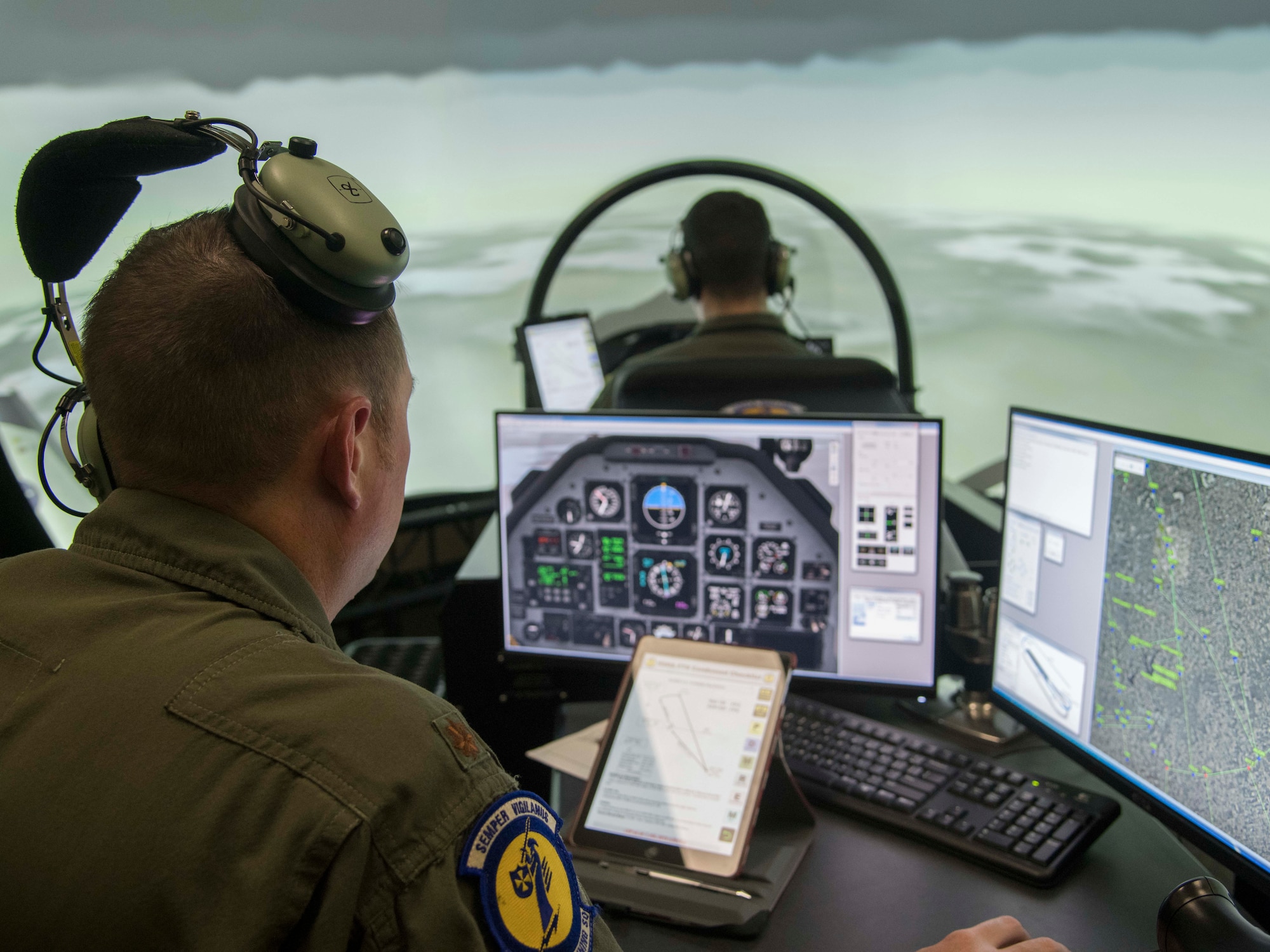 An instructor pilot sits at a console with two monitors that depict cockpit instruments on the left screen and a map of Randolph Air Force Base on the right. Directly infront of him is a simulator cockpit occupied by a student pilot.
