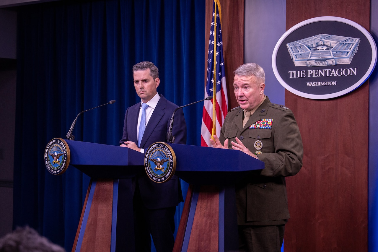A Marine Corps general speaks at a lectern as a civilian at an identical lectern stands beside him.