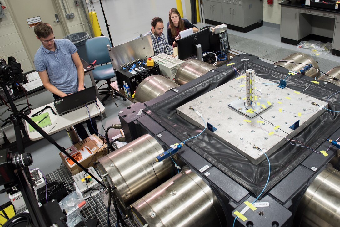 In a laboratory setting, three civilians provide computer inputs to a large mechanical apparatus.