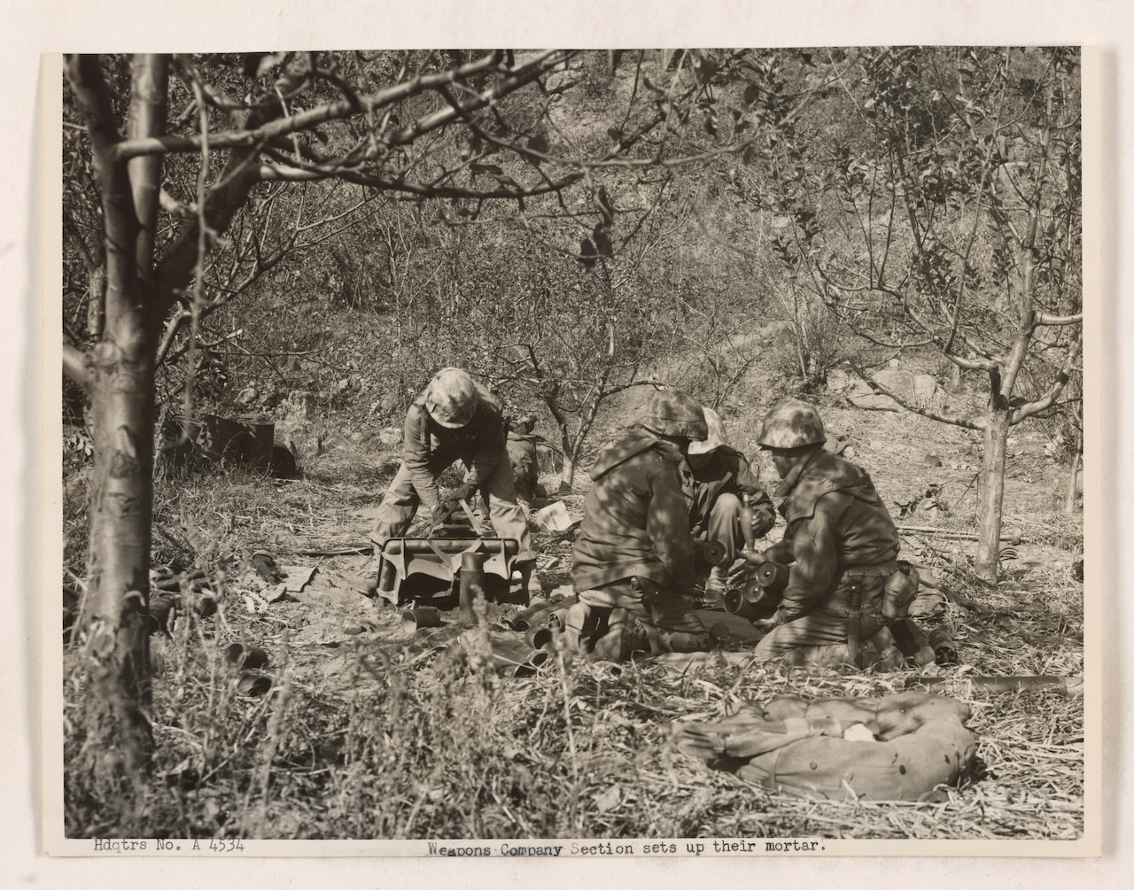 Four Marines work to put together a large gun while surrounded by brush and leafless trees.