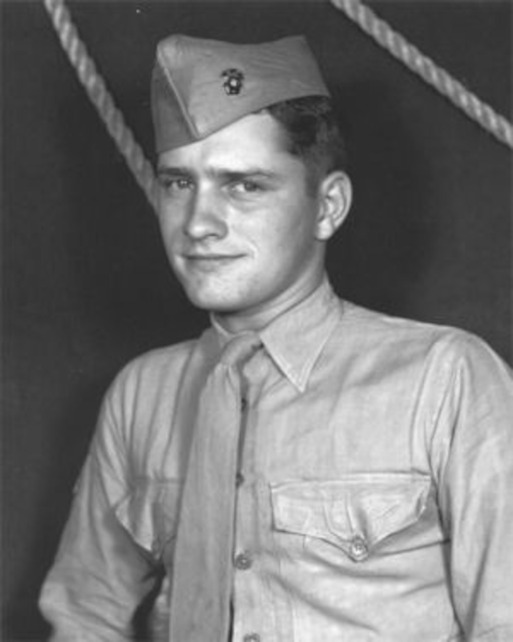 A young man wearing a shirt, tie and cap poses for a photo. Two ropes can be seen behind him.