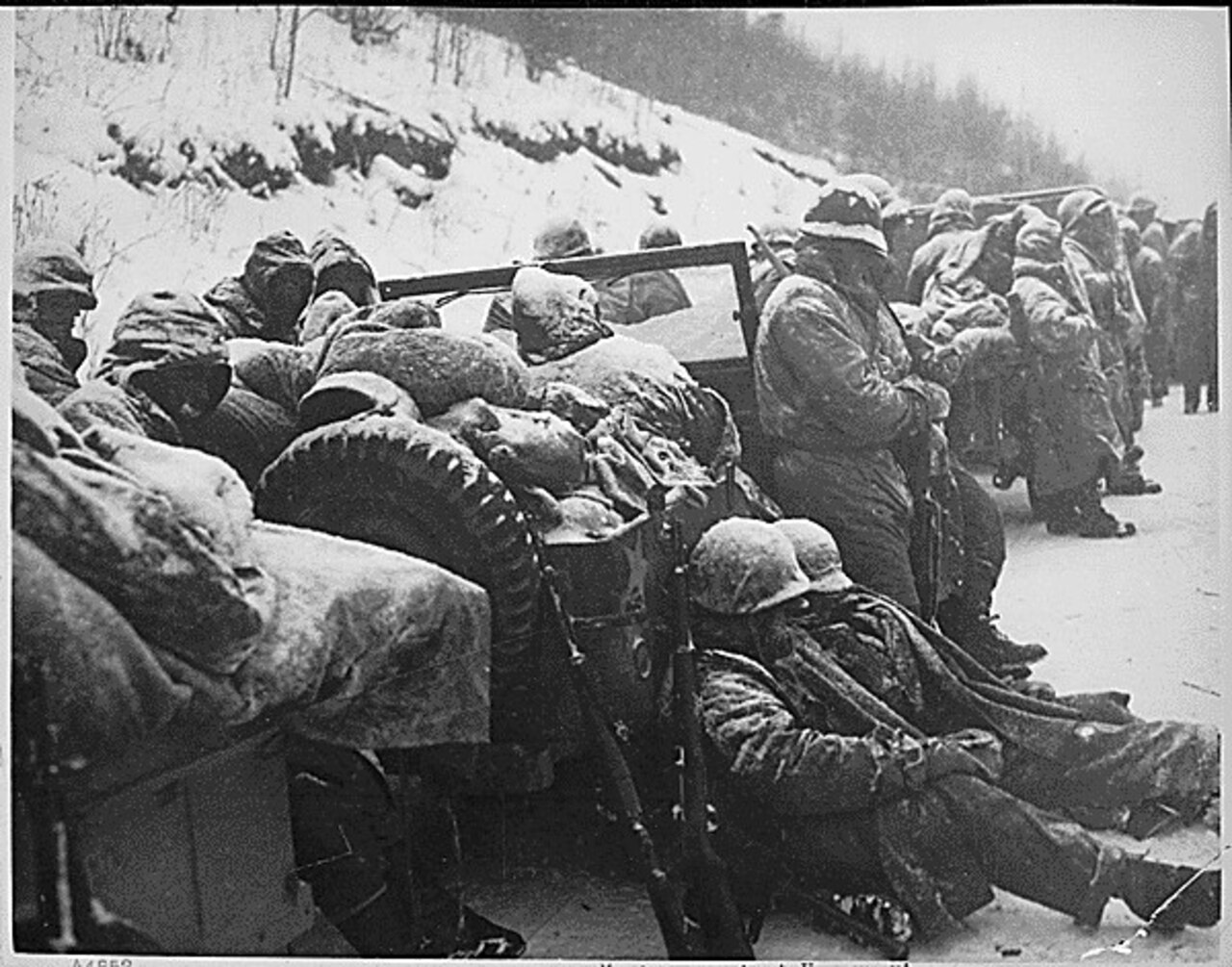 Marines in winter combat gear hold rifles and wait by trucks in the snow by a hill.
