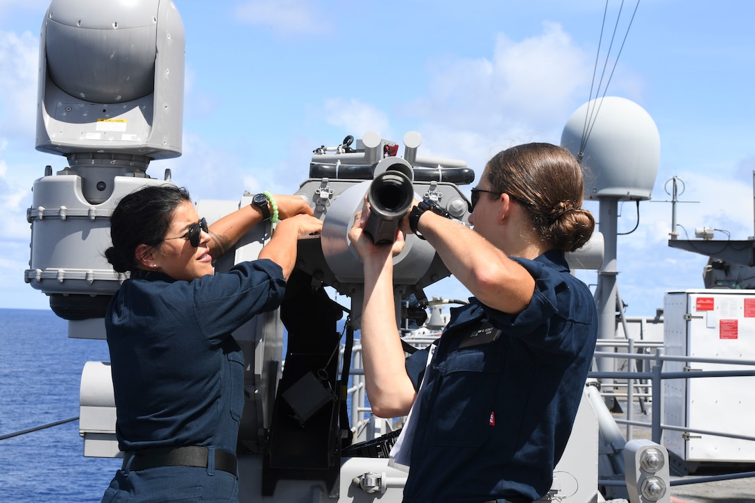 Two sailors remove the barrel of a machine gun on a military ship.