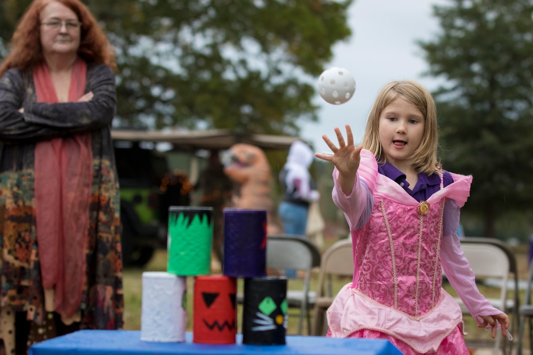 A child throws a ball at tin cans as a woman stands behind.