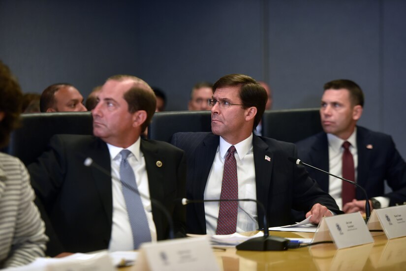 Three men in suits sit at a table, looking to their right, during a briefing.