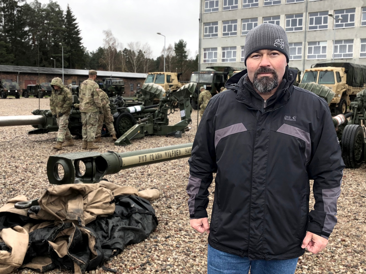 Photographic illustration of Customer Support Representative in front of military personnel preparing large scale equipment in a gravel parking lot.
