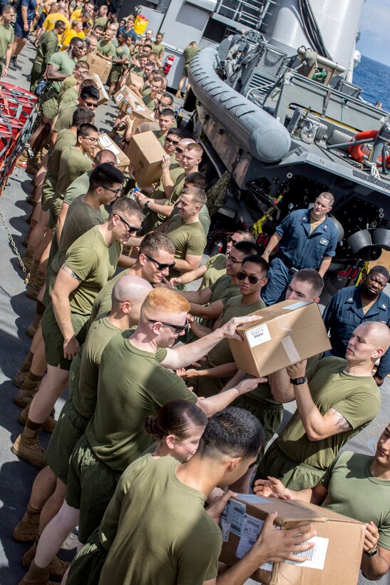 Vertical image of Marines tasing boxes of supplies onboard the USS Harpers Ferry.