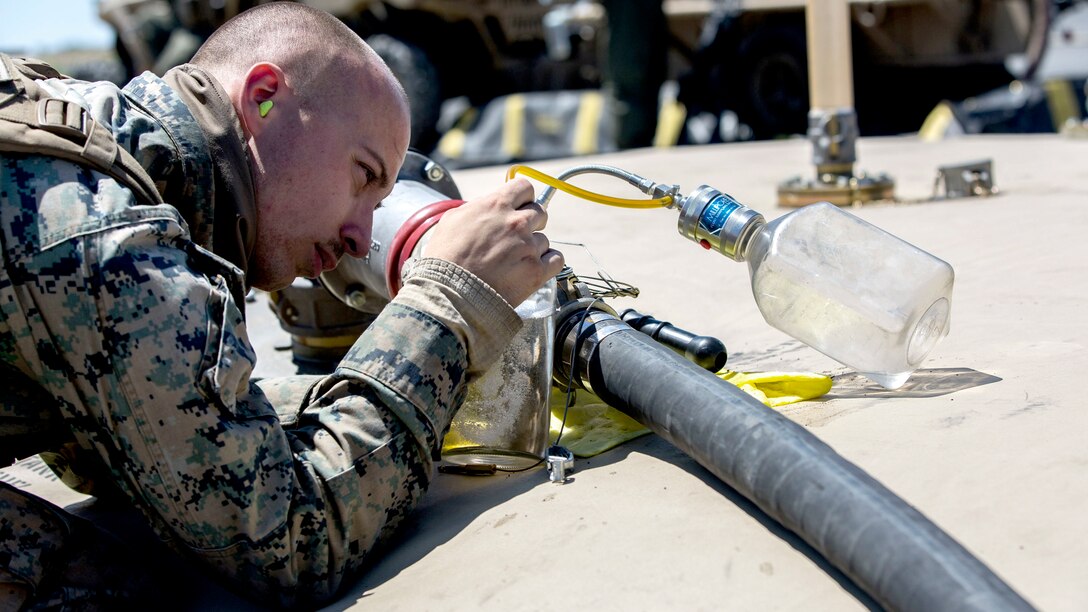 Image of Marine placing tubes connected to a glass jar into a bottle, with a fuel hose on the ground nearby.
