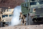 Image of a Marine exiting a green cammo truck with a smoke cloud behind it and a Humvee in the background.