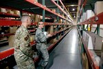 Image of two Air Force personnel with large warehouse shelving units extending back to the warehouse wall.