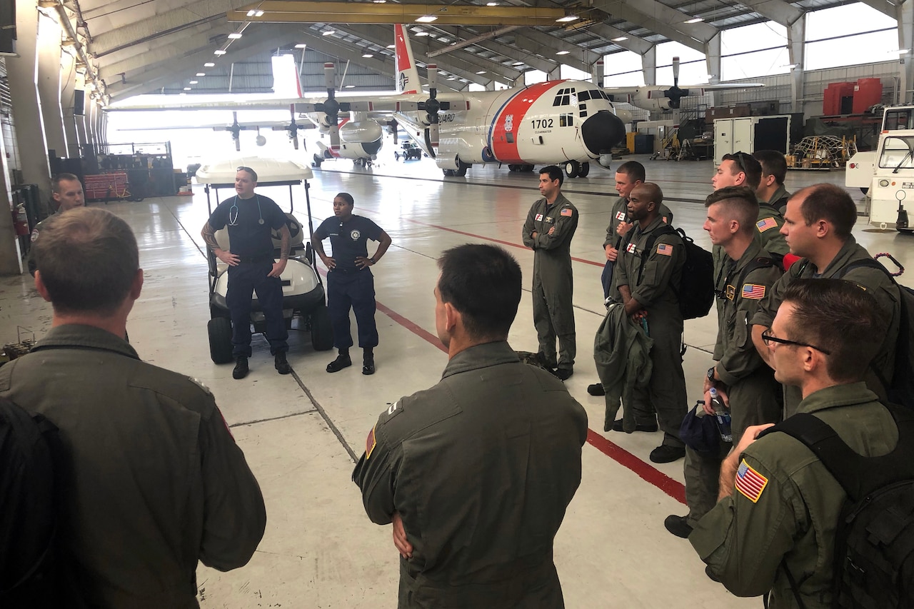 A group of service members listen to a briefing in an airplane hangar.