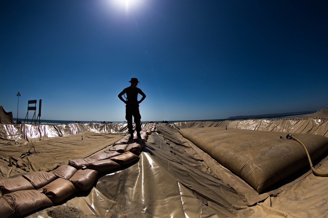 Image of a fuel pit with sandbags for walking, silhouette of Marine standing with fuel bladder being filled to the right, deep blue sky above.
