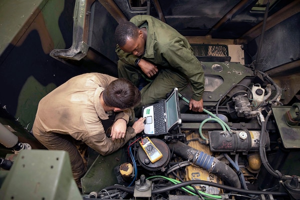 Image of two mechanics in Medium Tactical Vehicle Replacement compartment using a computer with multiple tubes and hoses and electronic meter system.
