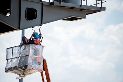 Image of two sailors in a lift bucket painting the underside of an extended part of a ship's hull.