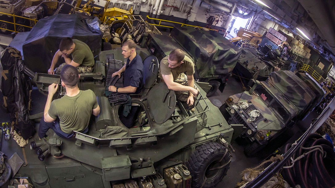 Image of four Marines sitting on top of a light armored vehicle, with other vehicles extending to the back of the ship's hold compartment.
