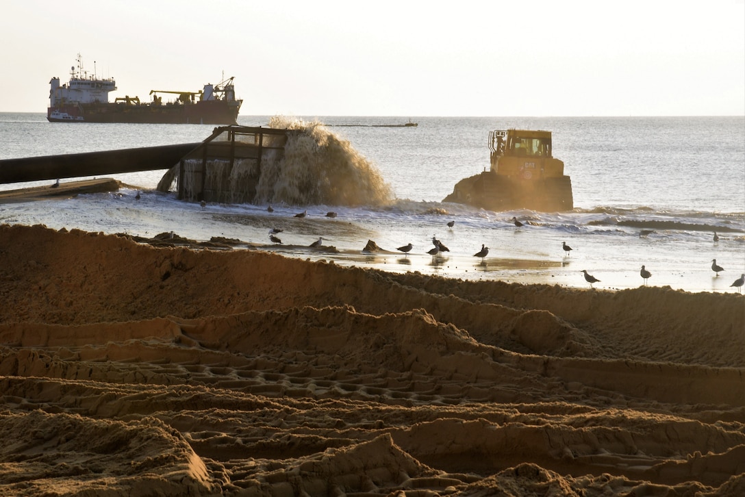 A pump and bulldozer move slurry around the shore as a hopper dredge is in the background during a beach renourishment operation