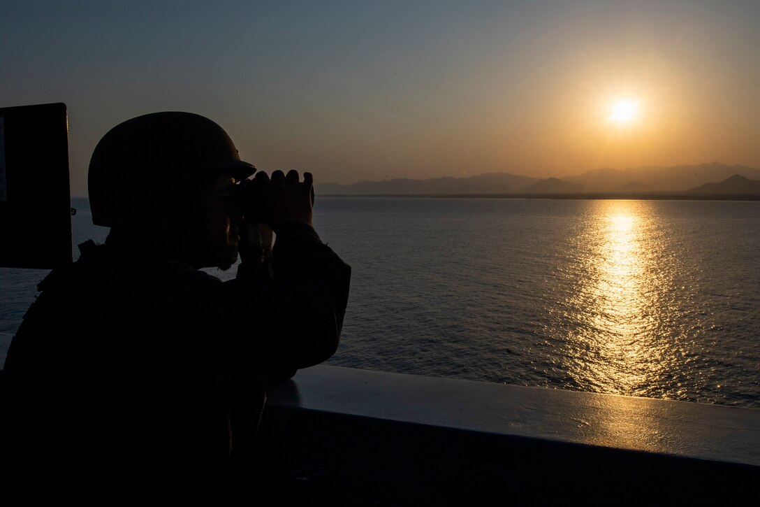 A silhouette of a sailor on a ship looking through binoculars at twilight.