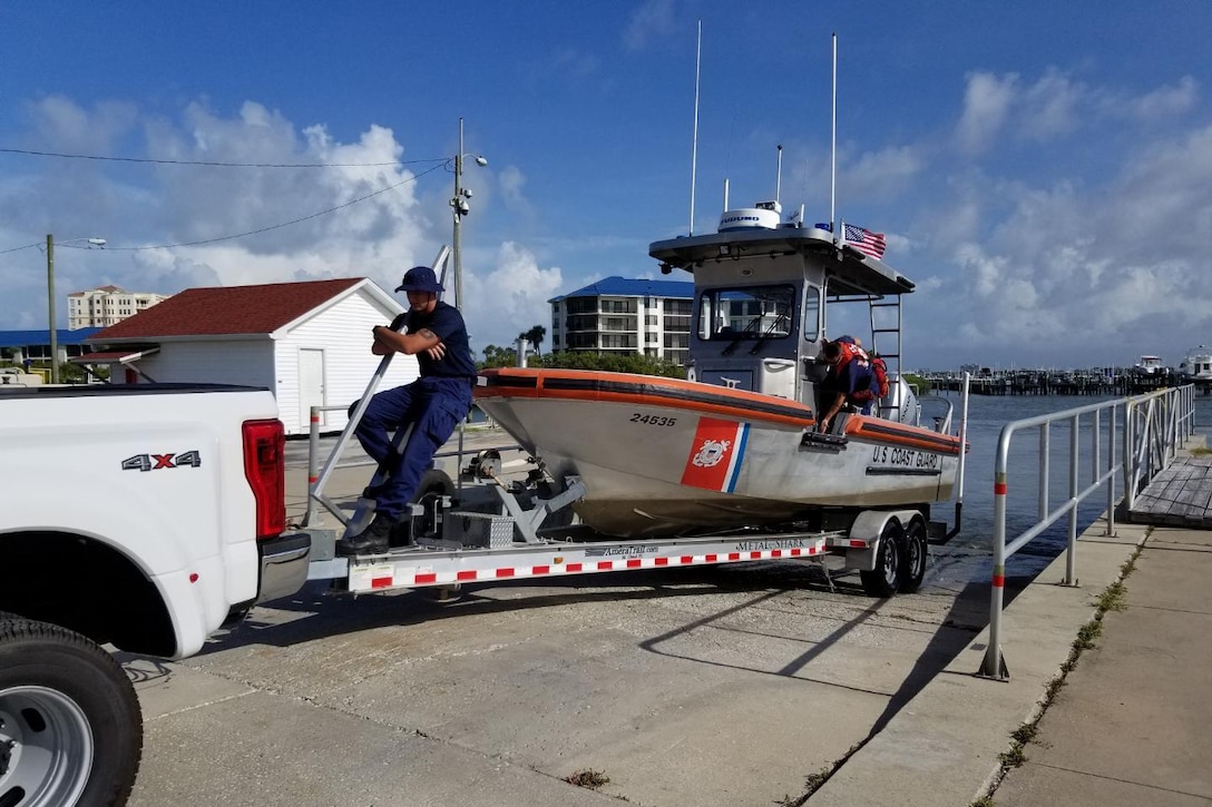 A Coast Guardsman rides on the bow of a boat being towed by a pickup truck.