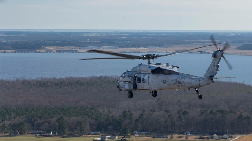A helicopter flies over rural terrain. In the far background is a large body of water.