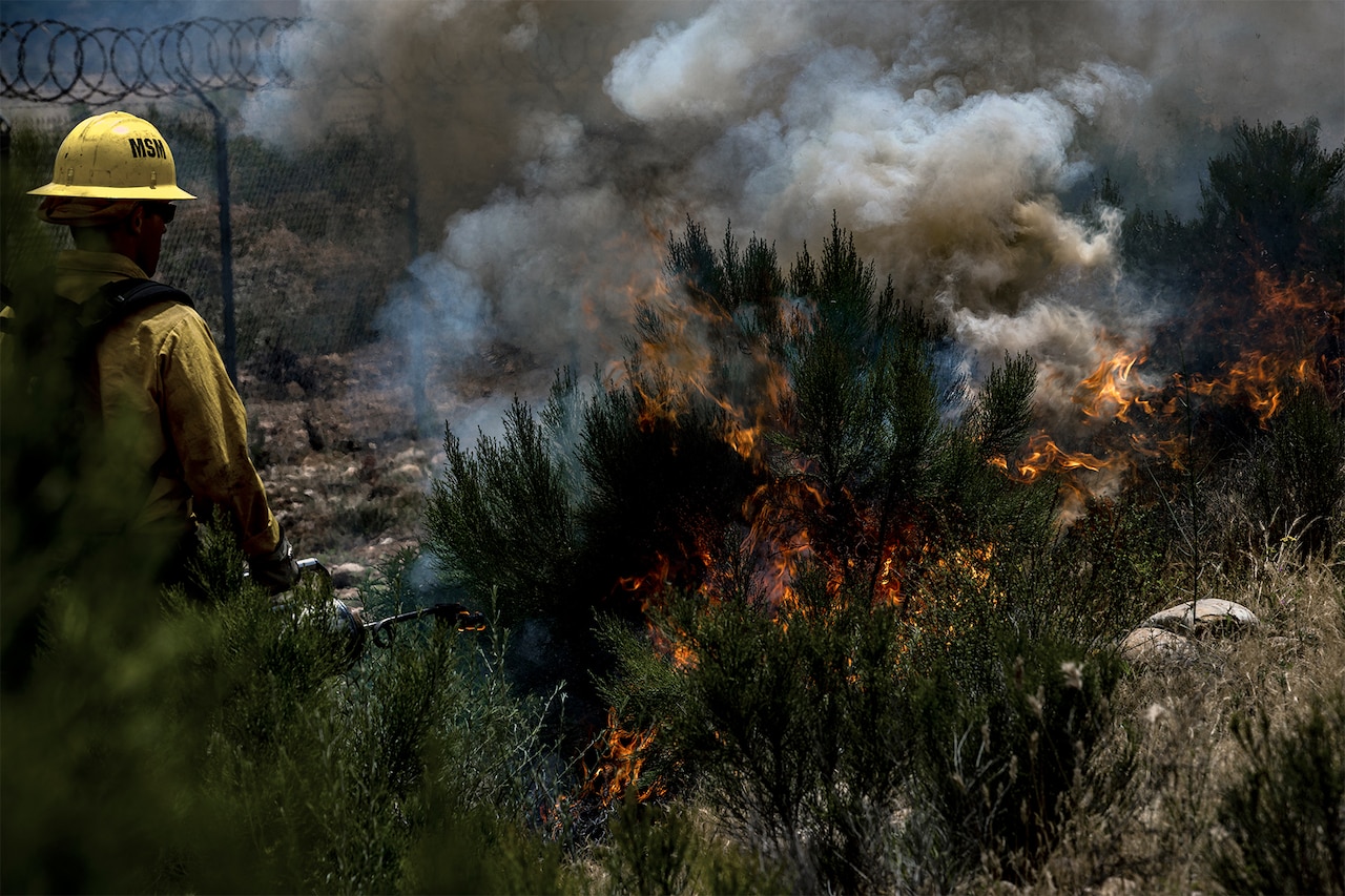 A firefighter wearing a yellow uniform watches over bushes and other vegetation as it burns. Thick smoke rises from the flames. To the left, concertina wire tops a fence.
