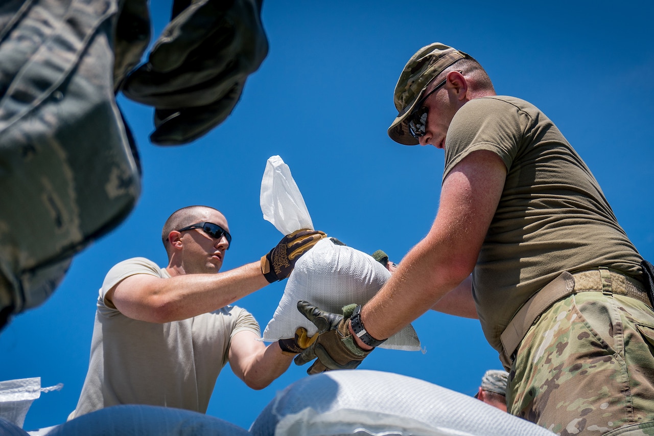 Looking upward towards a clear, blue sky, service members pass sandbags to one another.